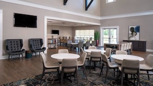 A spacious room with multiple seating areas, including round tables and chairs, a television mounted on the wall, and a bookshelf.