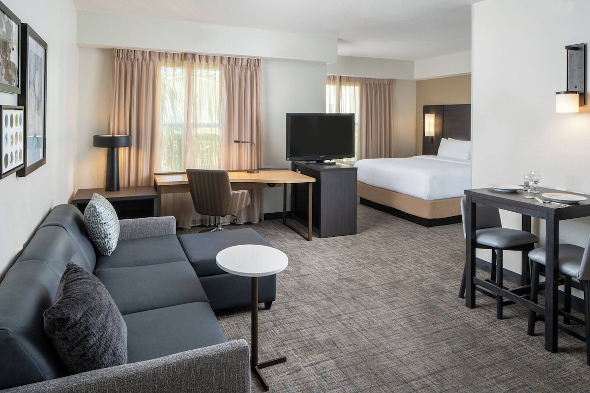 A hotel room with a sofa, a small round table, a desk with a TV, a bed, and a window with curtains.