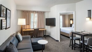 A hotel room with a sofa, a small round table, a desk with a chair, a bed, a TV, and a mirror.