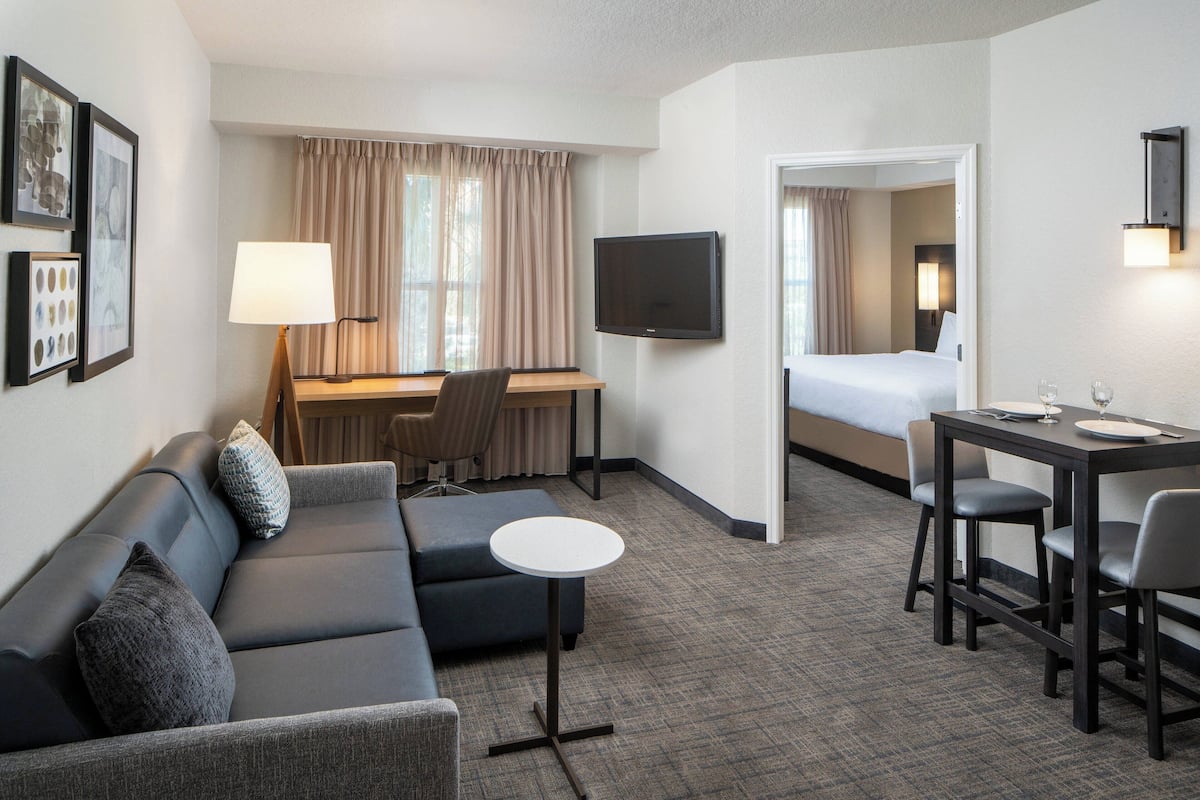 A hotel room with a sofa, a small round table, a desk with a chair, a bed, a TV, and a mirror.
