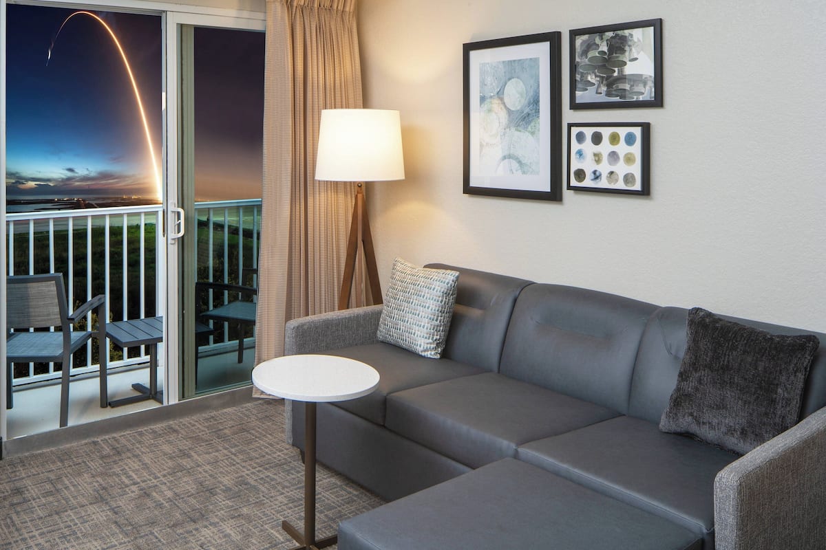 A modern living room with a grey sofa, a small round table, and a lamp. There are two framed artworks on the wall and a balcony with a view of the sky.