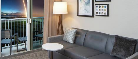 A modern living room with a grey sofa, a small round table, and a lamp. There are two framed artworks on the wall and a balcony with a view of the sky.