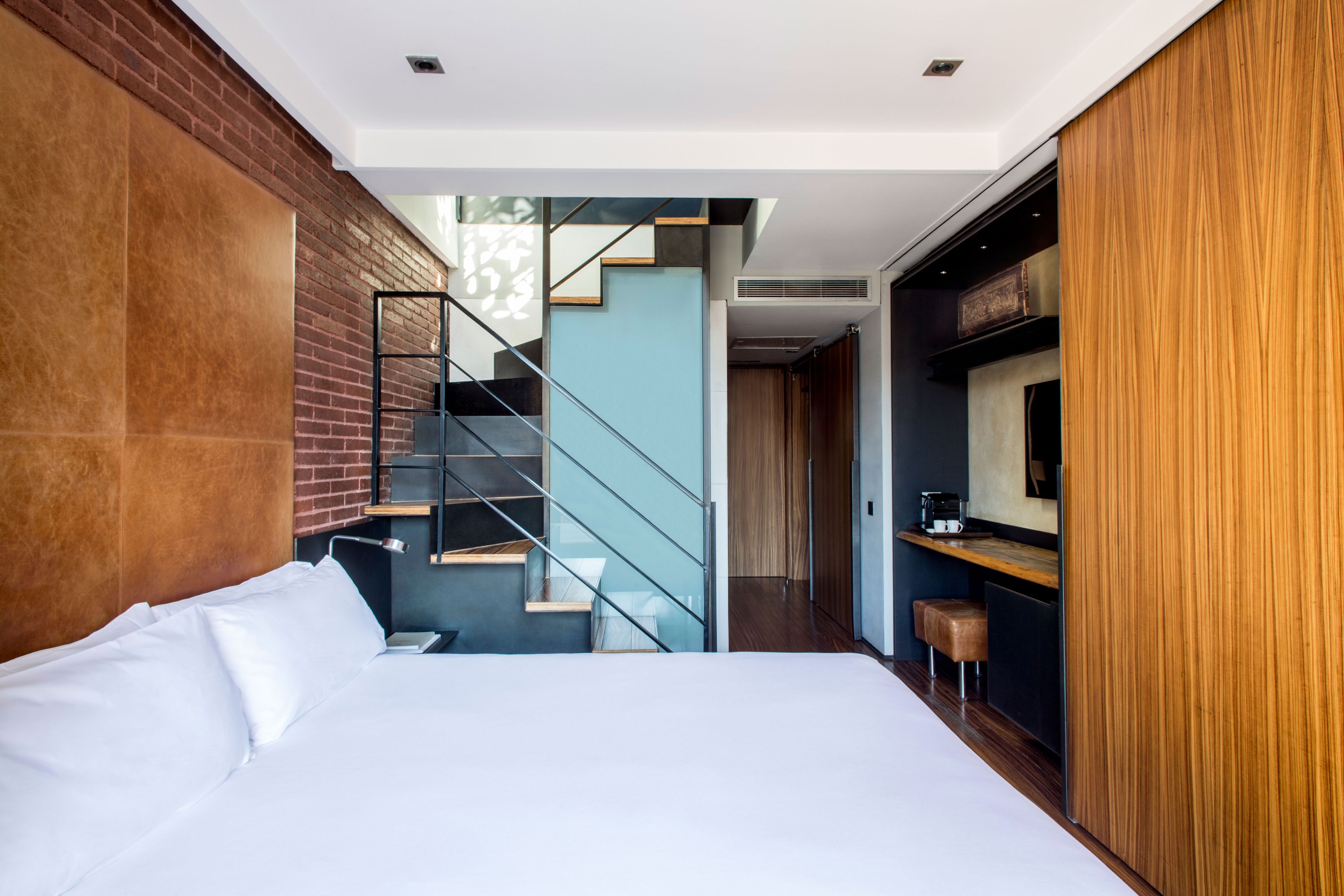 Una habitación de hotel moderna con una cama grande, una pared decorada con ladrillos, una escalera que lleva a una zona superior y un armario de madera.