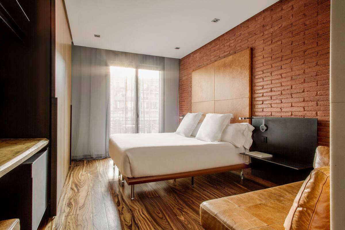 Une chambre d’hôtel moderne avec un plancher en bois, un grand lit avec du linge de lit blanc, un mur décoratif en briques et un fauteuil en cuir.