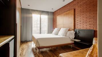 Una habitación de hotel moderna con piso de madera, una cama grande con ropa blanca, una pared de acento de ladrillo y un sillón de cuero.
