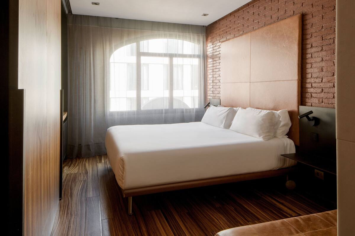 Une chambre d’hôtel moderne avec un grand lit, un mur décoratif en briques et une grande fenêtre agrémentée de rideaux légers.