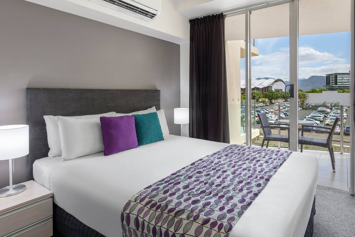 A hotel room with a large bed, a nightstand, a lamp, a window with a view of a parking area and mountains.