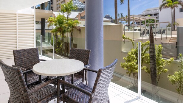 Un balcón con mobiliario de mimbre, una mesa de vidrio y vistas a palmeras y edificios.