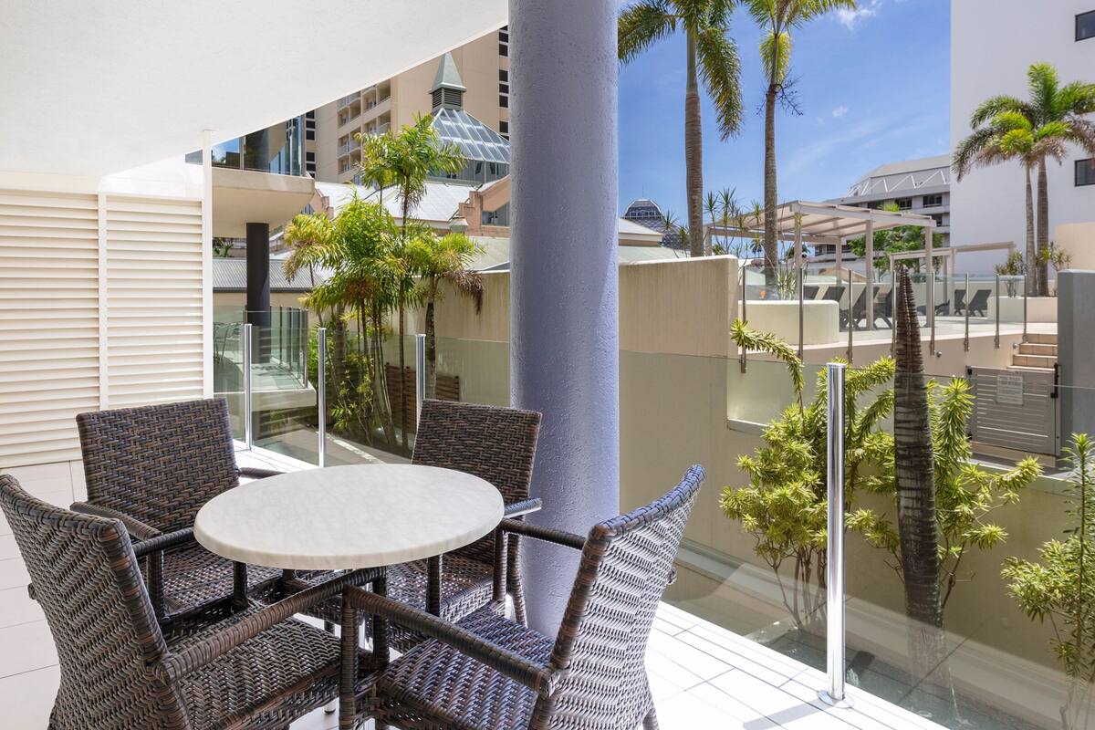 A balcony with wicker furniture, a glass table, and a view of palm trees and buildings.
