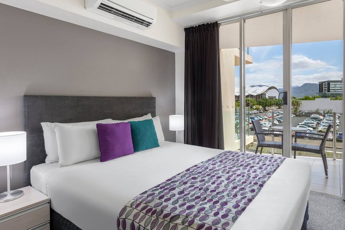 A hotel room with a bed, a nightstand, a lamp, a window with a view of a parking lot and mountains.