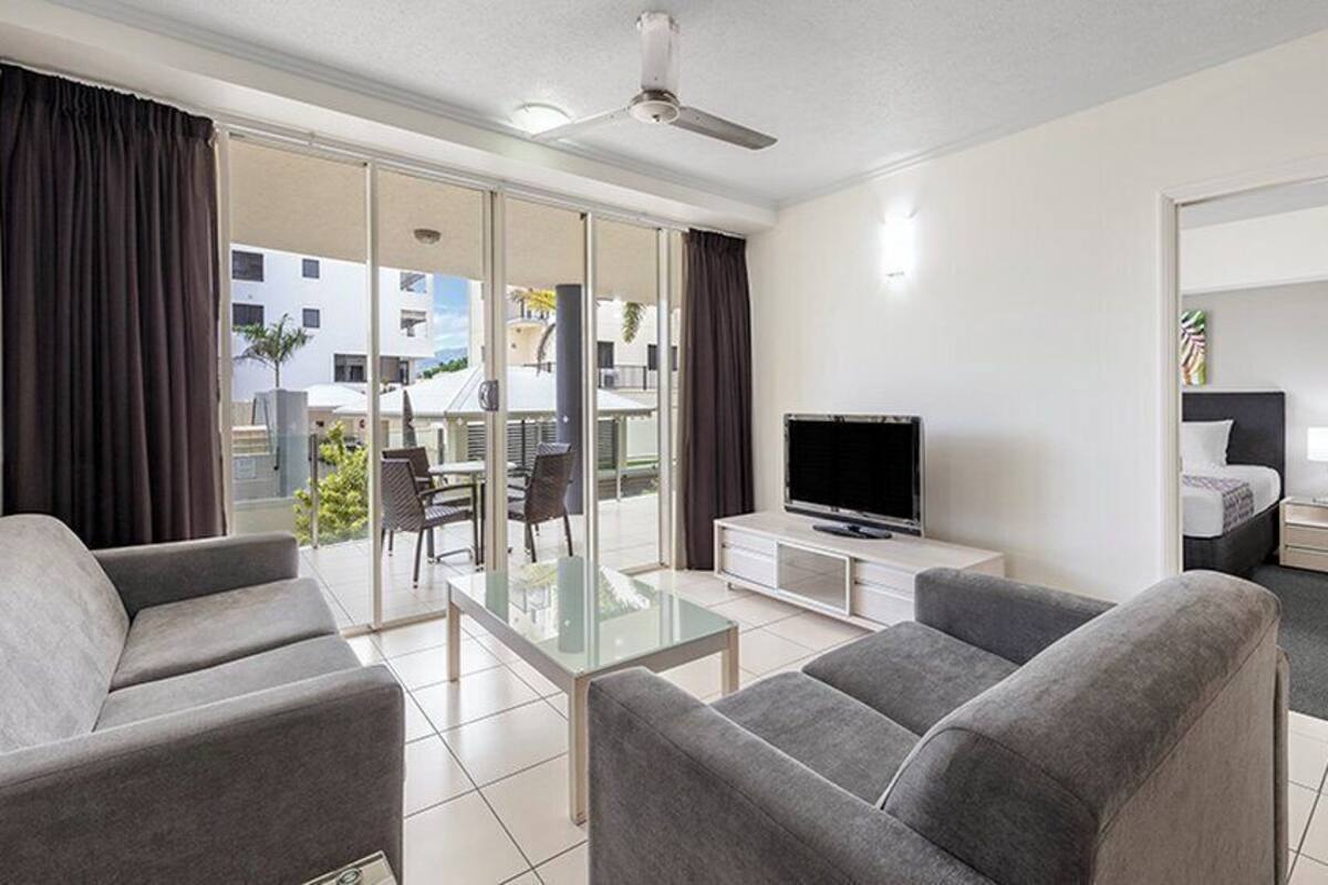 A modern hotel room with a flat-screen TV, grey sofas, a glass coffee table, and a balcony with a view of the city.