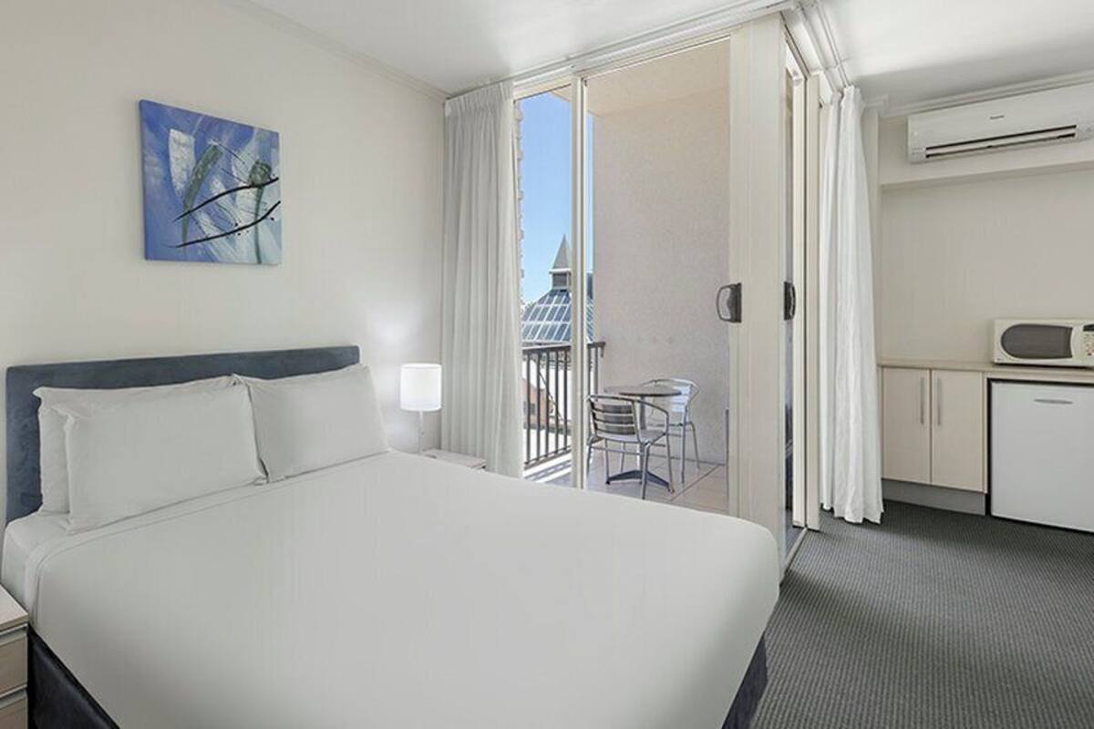 Habitación de hotel con una cama grande, un escritorio pequeño con microondas, una silla y un balcón con vistas a edificios.