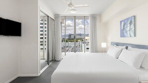 Habitación de hotel con una cama grande, ventilador de techo, televisor montado en la pared y vista a un estacionamiento y edificios.