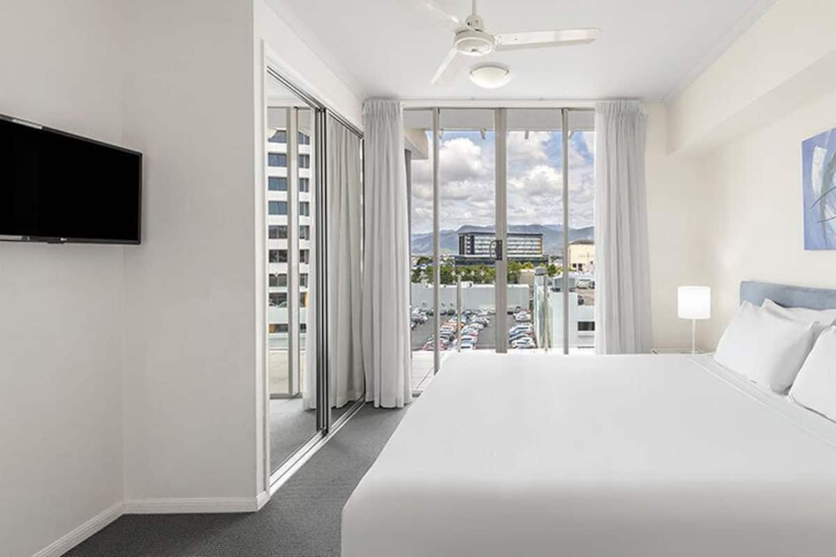 Habitación de hotel con una cama grande, ventilador de techo, televisor montado en la pared y vista a un estacionamiento y edificios.
