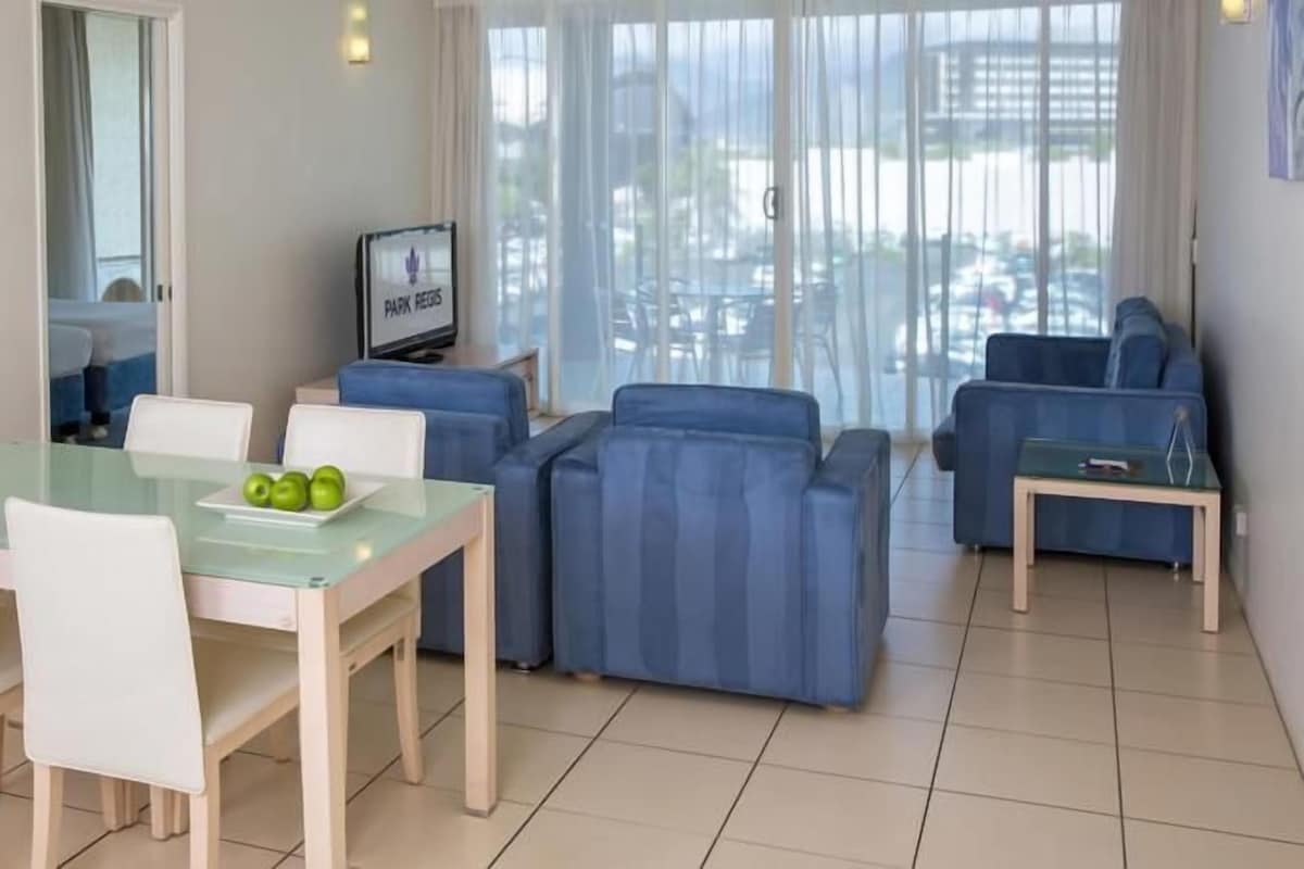 Una sala de estar moderna con comedor, que incluye una mesa de vidrio, sofás azules y un televisor.
