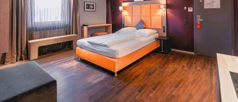 Habitación de hotel moderna con piso de madera, cama con sábanas blancas, escritorio, un banco y una puerta con cerradura roja.