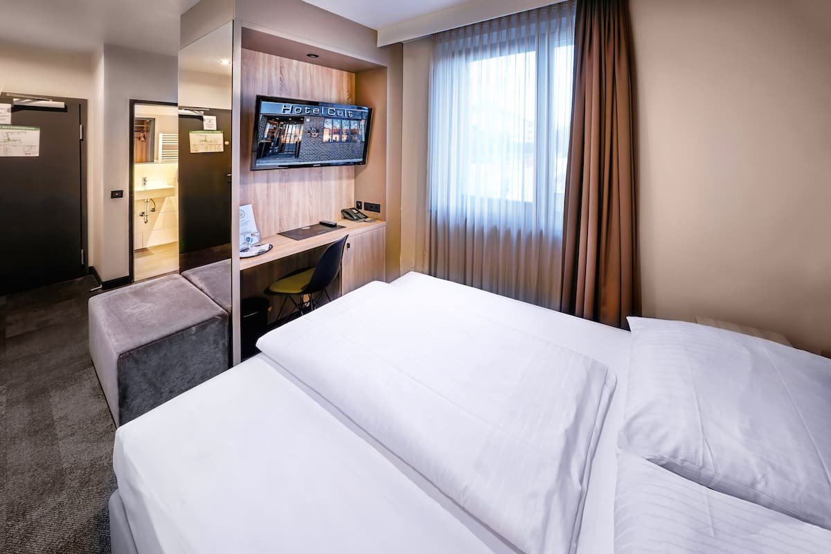 A hotel room with a bed, a desk with a computer, a TV, and a window with curtains.