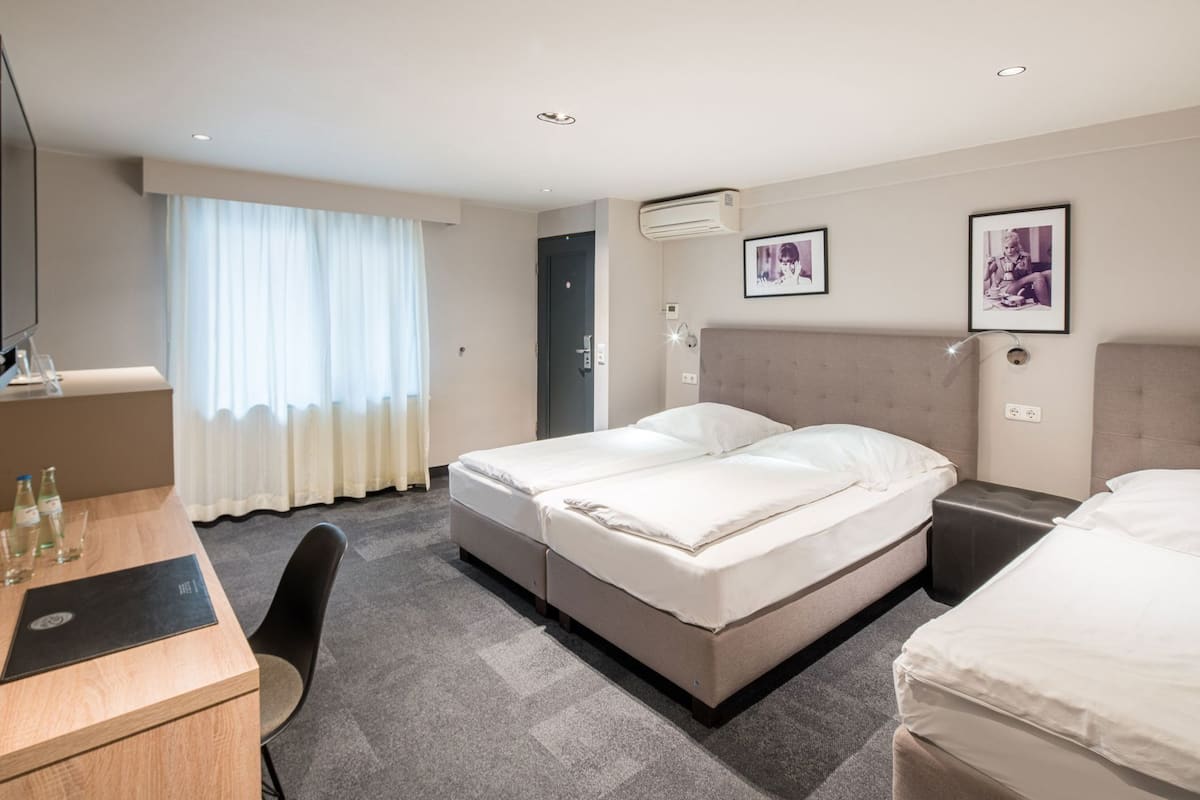 A hotel room with a bed, a desk, a chair, an air conditioning unit, and two framed pictures on the wall.
