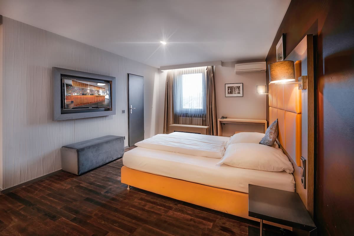 A modern hotel room with a large bed, a bench, a TV, and a window with curtains.