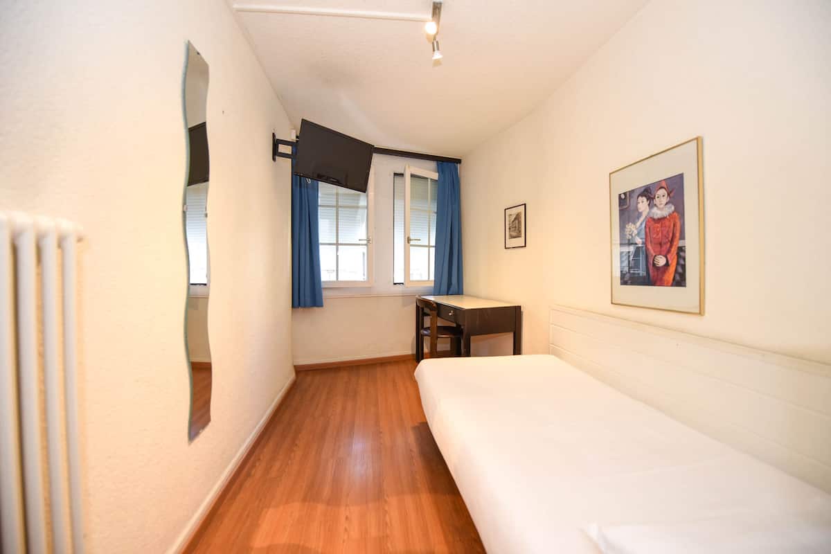 A room with a bed, a desk, and a chair. There is a window with curtains, a radiator, and a framed picture on the wall.