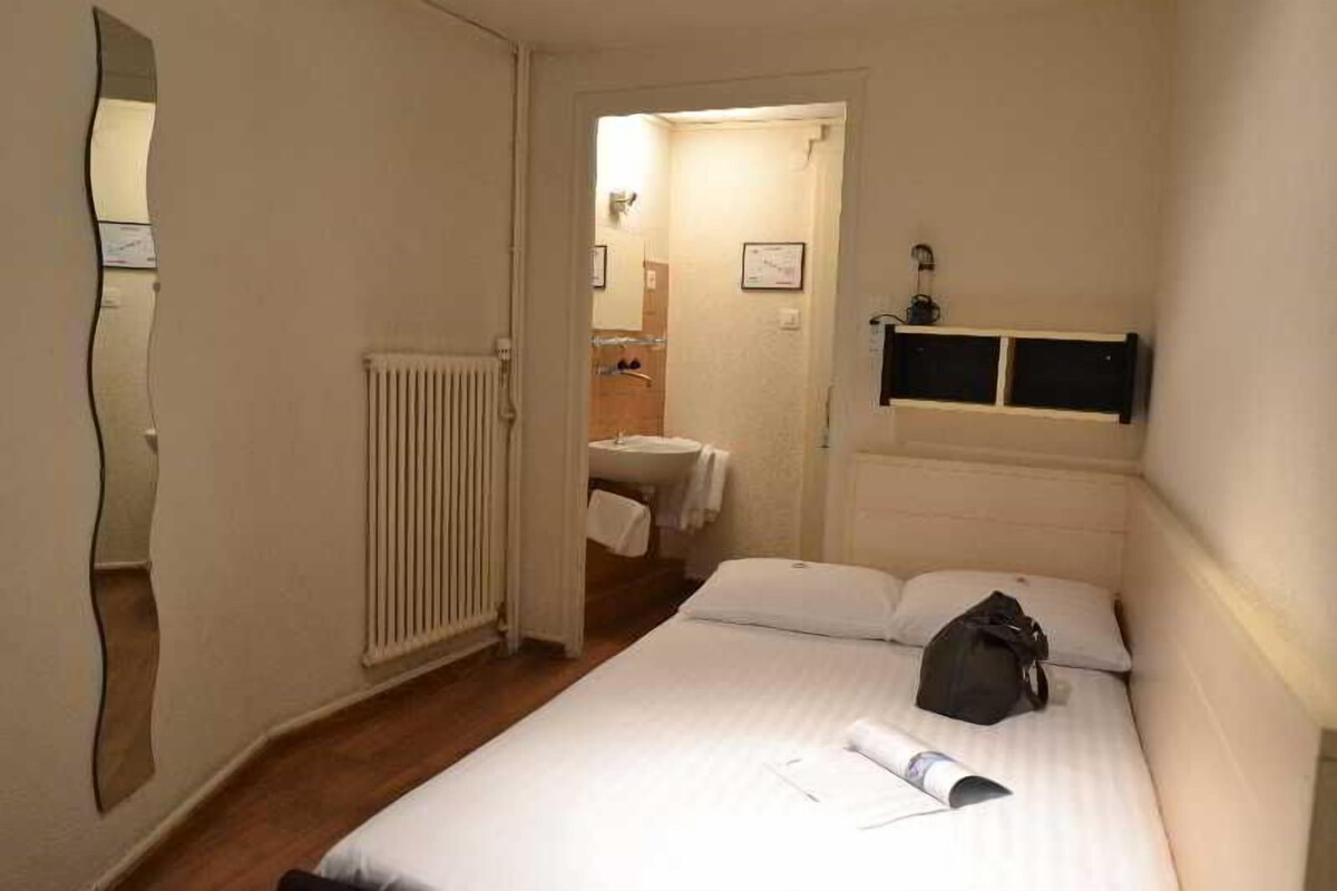 A hotel room with a bed, a radiator, a bathroom visible through an open door, and a shelf with a mirror.