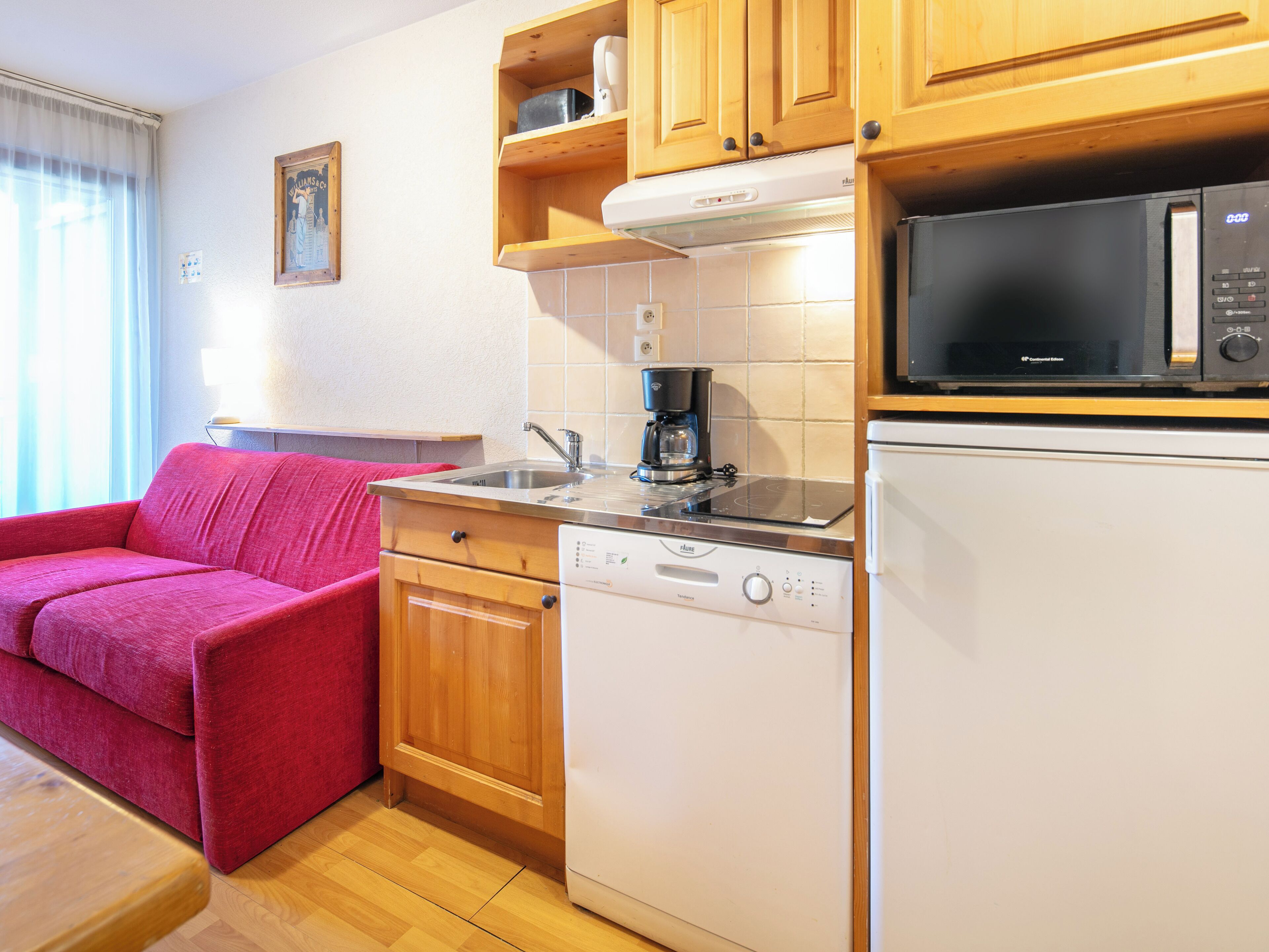 Una cocina compacta con armarios de madera, microondas integrado, lavavajillas y un sofá rojo.
