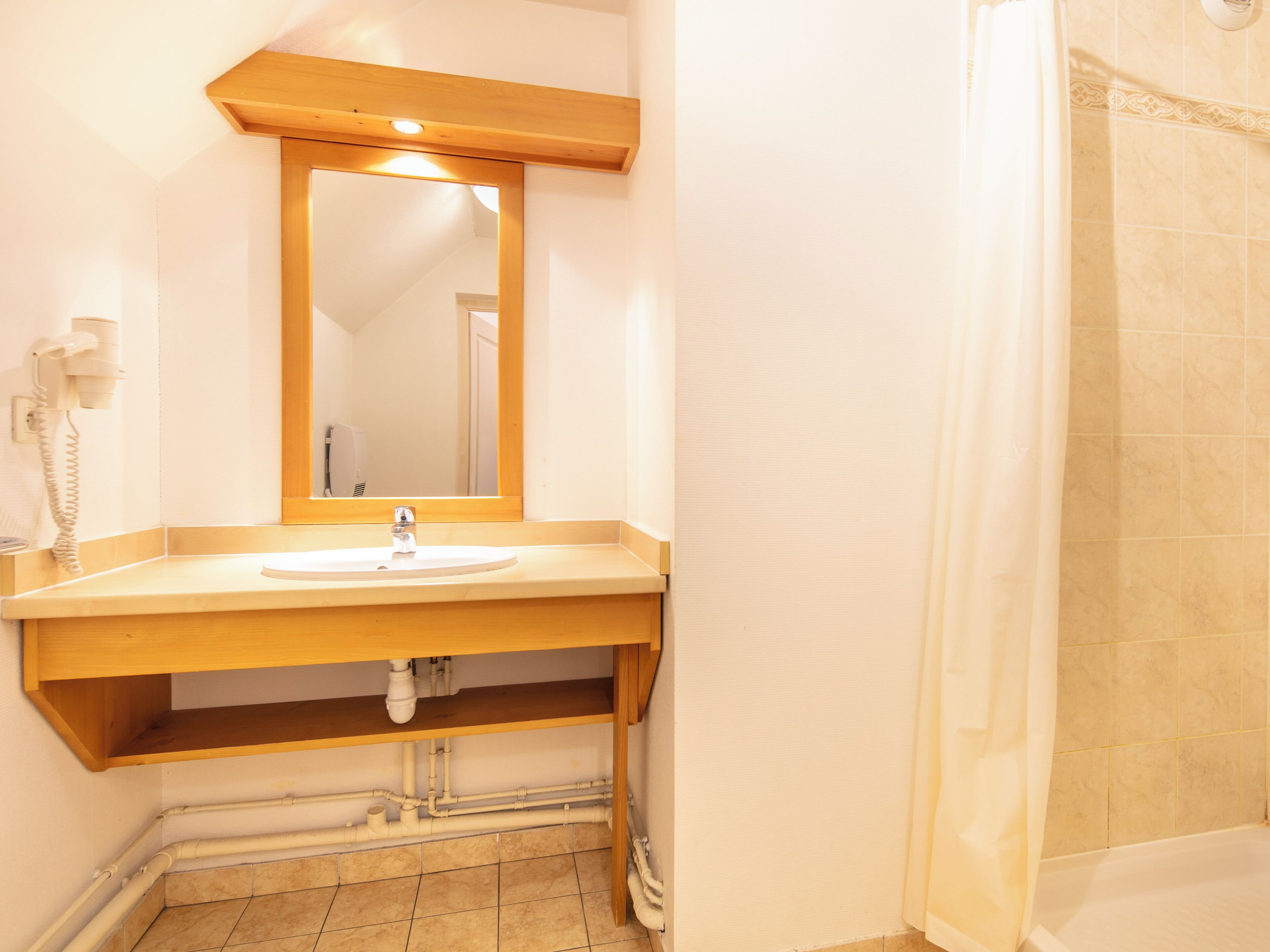 Un baño con tocador de madera, lavamanos, espejo, cortina de ducha y tuberías visibles.