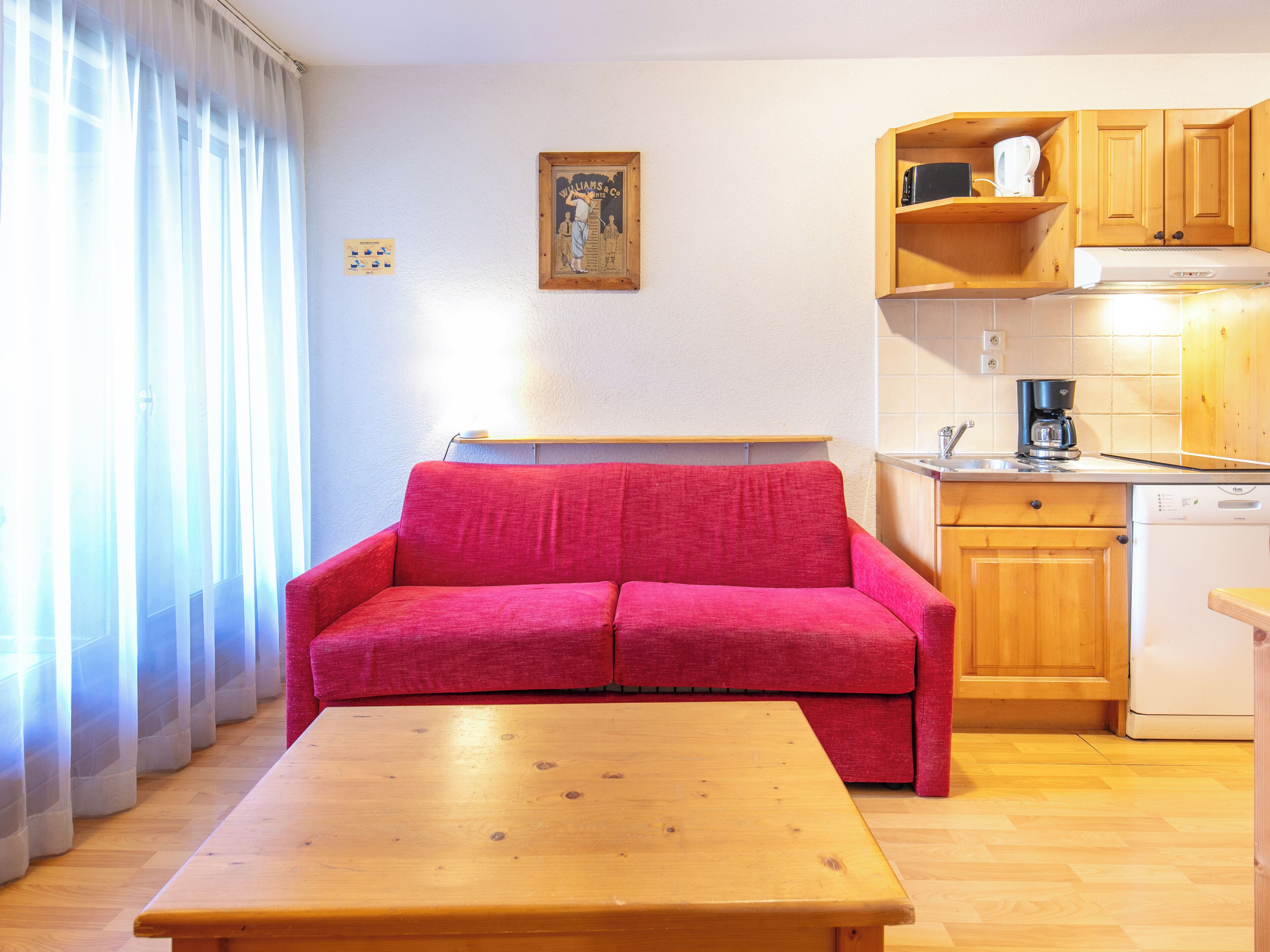 A compact living space with a red sofa, wooden table, kitchen area with wooden cabinets, and a window with sheer curtains.