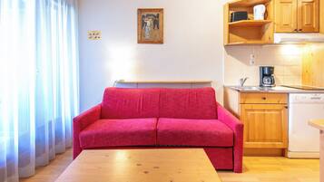 A compact living space with a red sofa, wooden table, kitchen area with wooden cabinets, and a window with sheer curtains.