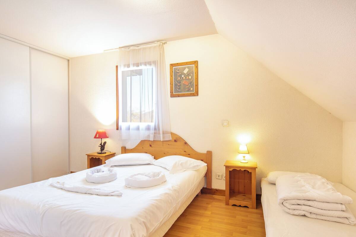 A bedroom with a single bed, a bedside table with a lamp, a window with sheer curtains, and a painting on the wall.