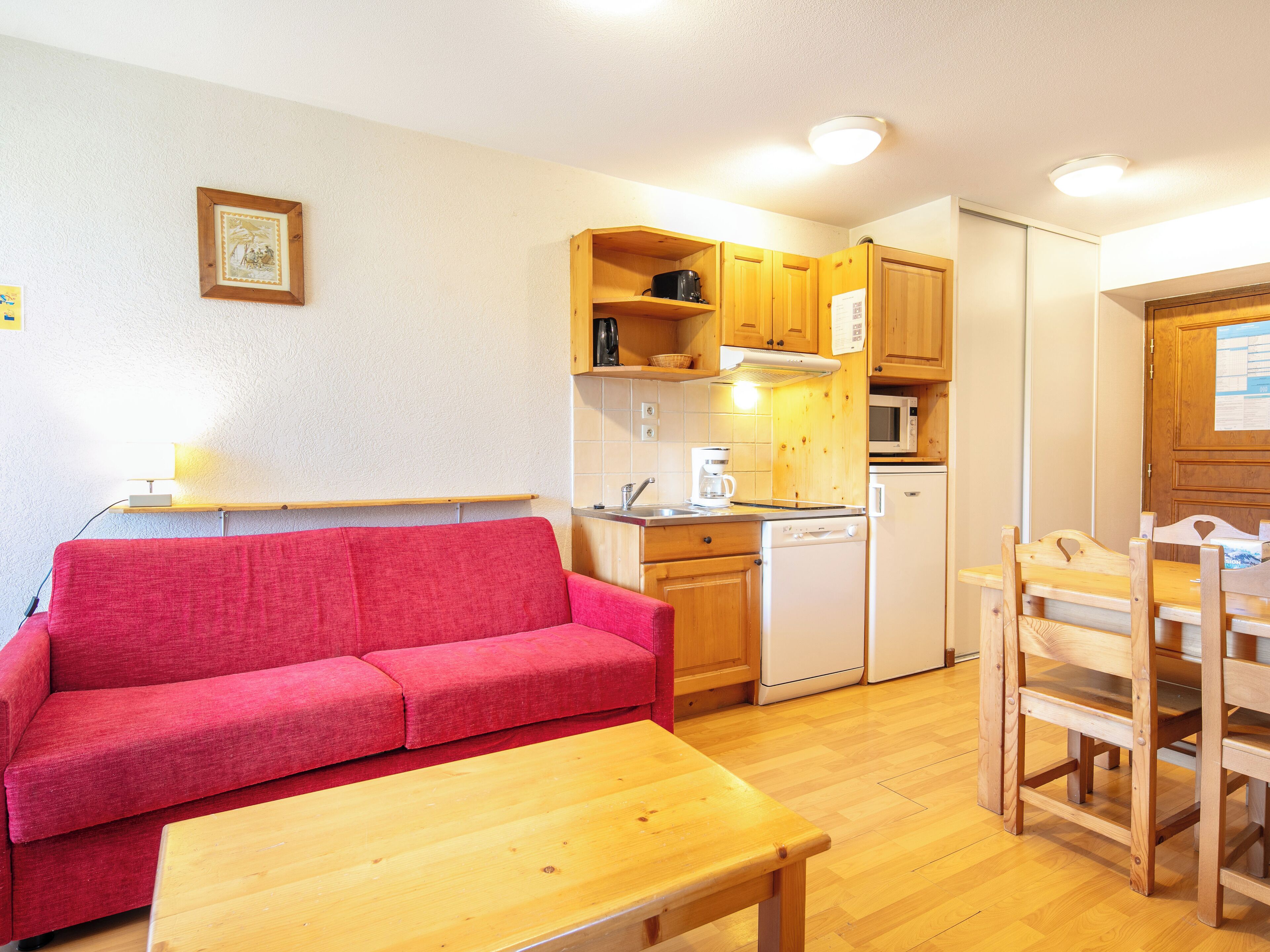 Un espace de vie compact comprenant un coin cuisine, un canapé rouge, une table en bois et un coin repas avec des chaises en bois.