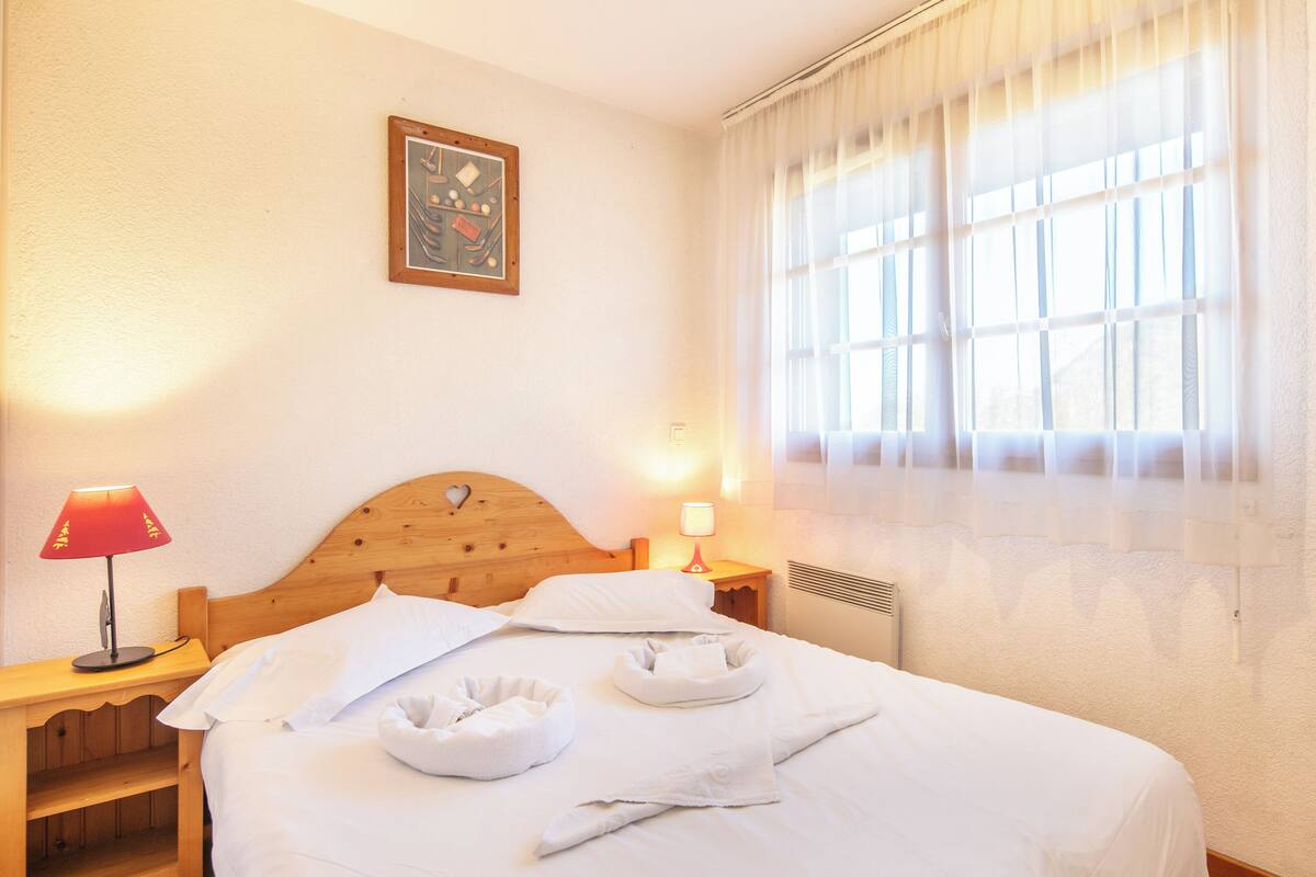 A neatly made bed with white linens, a wooden headboard, a bedside table with a lamp, and a window with sheer curtains.
