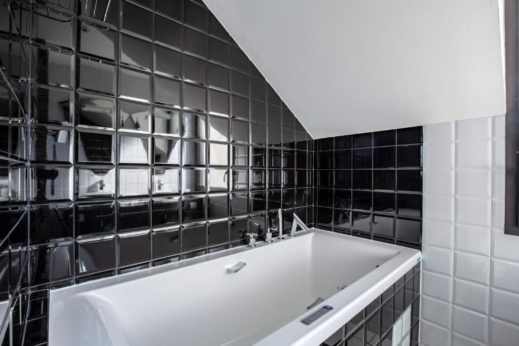 A modern bathroom with a large white bathtub, black and white tiled walls, and a visible stainless steel sink.