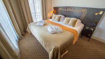 A hotel room with a large bed, a headboard with a decorative design, a bedside table with a lamp, and a window with curtains.
