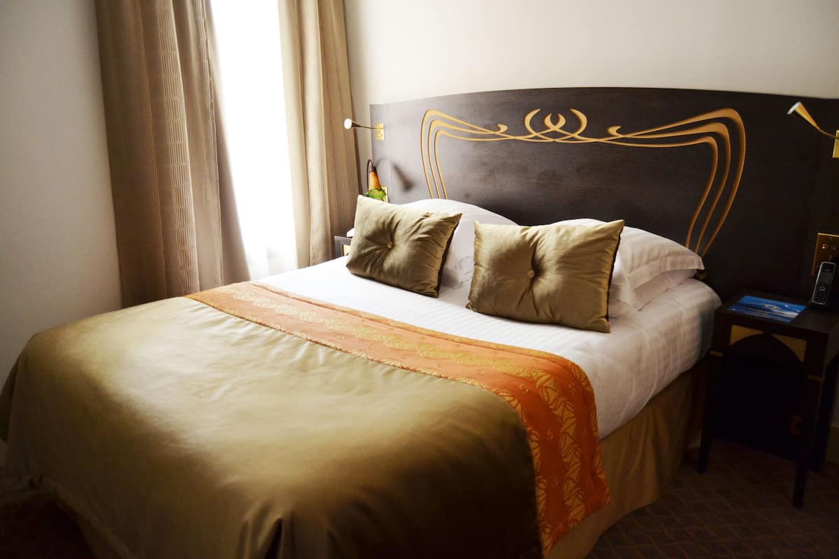 A neatly made bed with a gold and white bedspread, a decorative headboard, and a bedside table with a lamp.