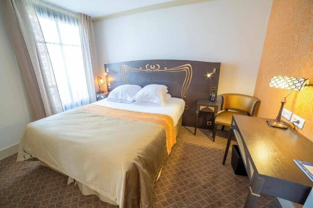 A hotel room with a large bed, a desk, a chair, and a nightstand with a lamp.