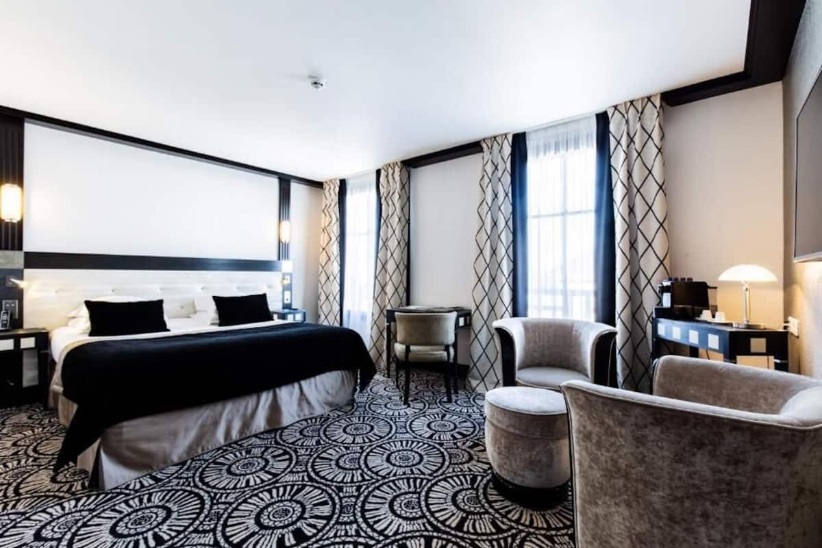 A hotel room with a large bed, two armchairs, a small table, and a patterned carpet.