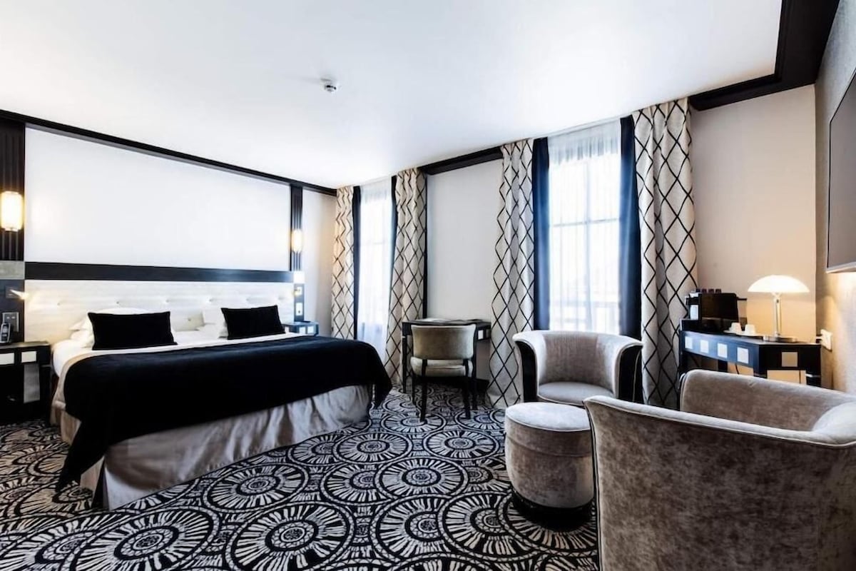 A hotel room with a large bed, two armchairs, a small table, and a patterned carpet.