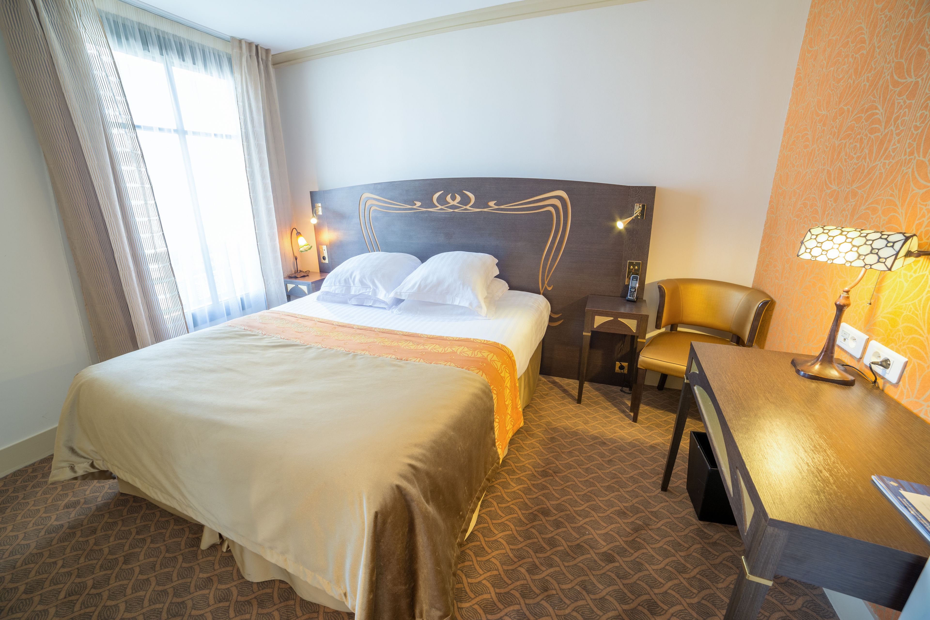A hotel room with a large bed, a desk, a chair, and a nightstand with a lamp.