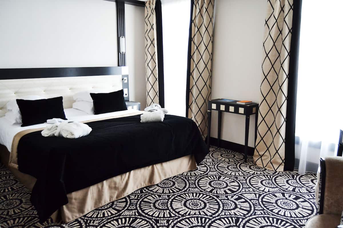 A hotel room with a large bed, black and white patterned carpet, a small side table, and a chair.