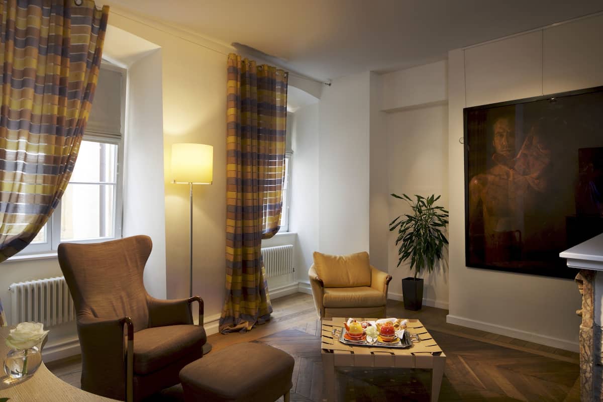 Un salon avec un fauteuil marron, un fauteuil jaune, une table basse sur laquelle se trouve un plateau de nourriture, une lampe sur pied et un tableau accroché au mur.