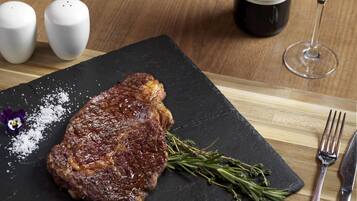 Un bistec sobre una tabla de pizarra con sal y romero, una botella de vino Aulo y saleros y pimenteros sobre una mesa de madera.
