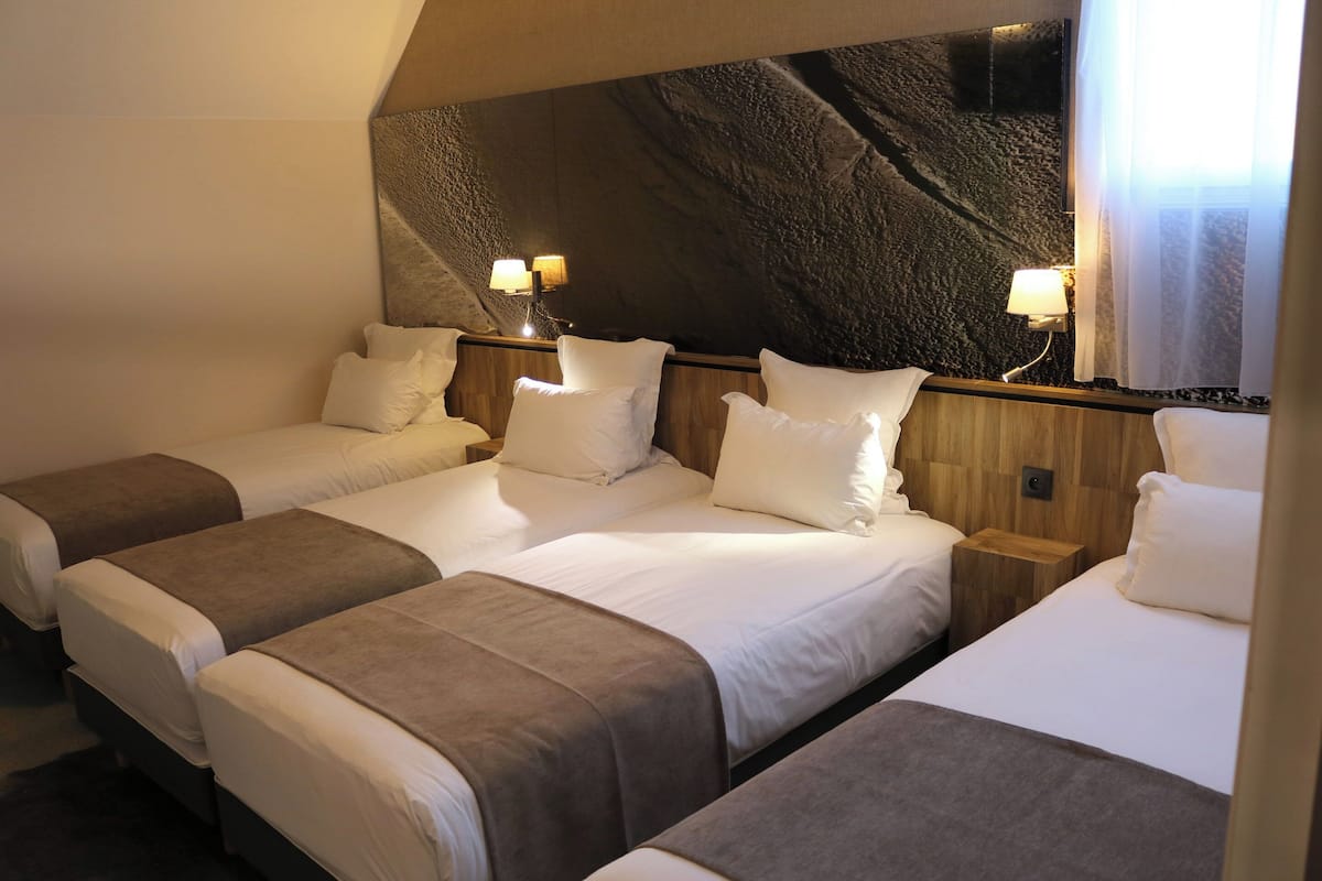 A hotel room with three beds, each with white linens and brown headboards. There are wall-mounted lamps on either side of the beds.