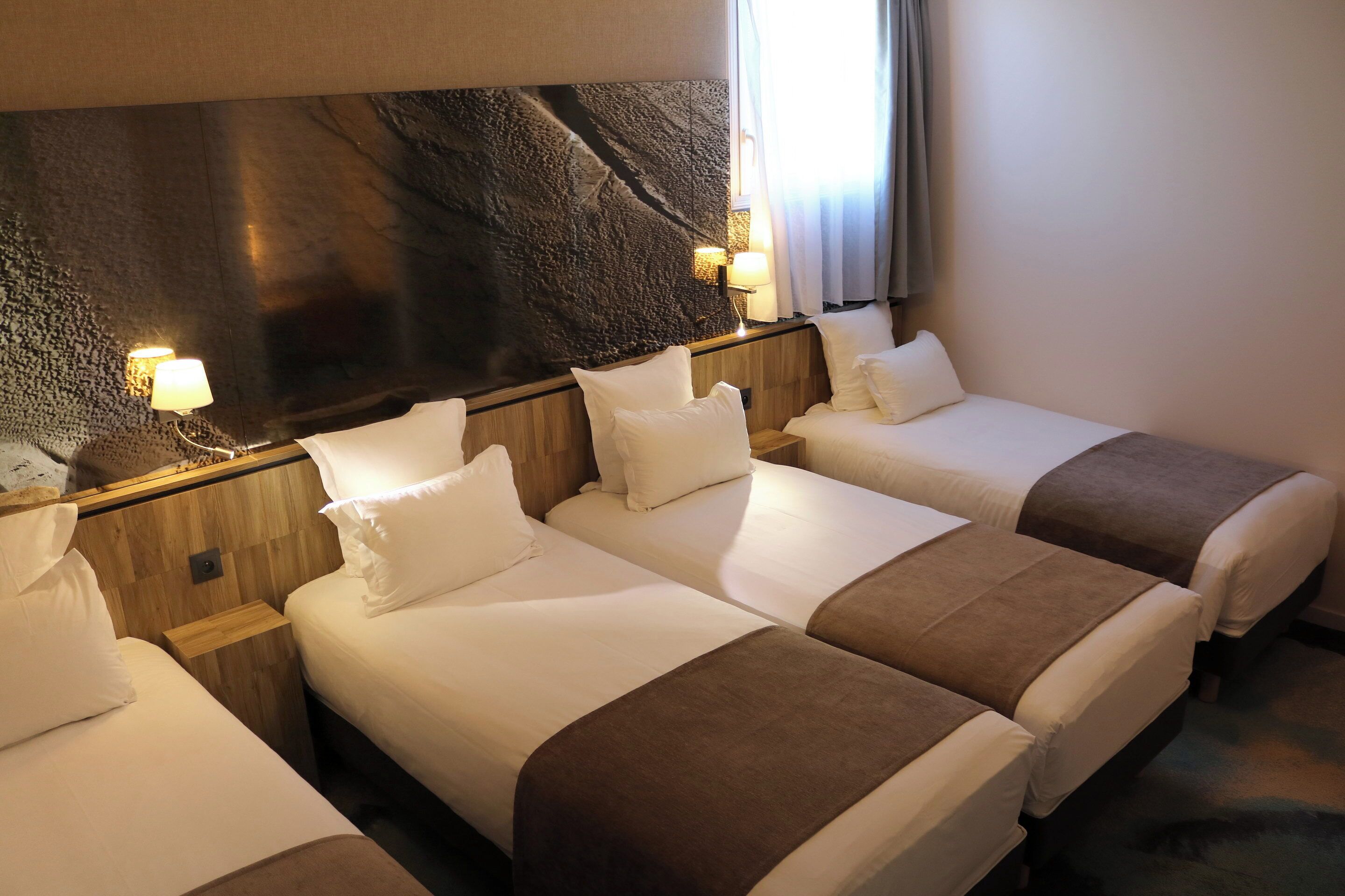 Une chambre d’hôtel avec quatre lits simples, une tête de lit en bois, des lampes fixées au mur et un grand mur à texture.