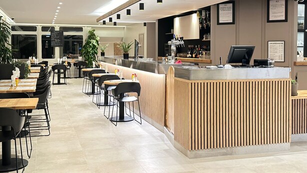 A modern cafe with a counter, tables, chairs, and potted plants. - Grand Hôtel Senia (Orly)