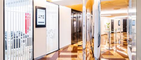Un couloir d’hôtel moderne avec un comptoir d’enregistrement, un espace de réception et une cloison en verre ornée d’une photographie encadrée.
