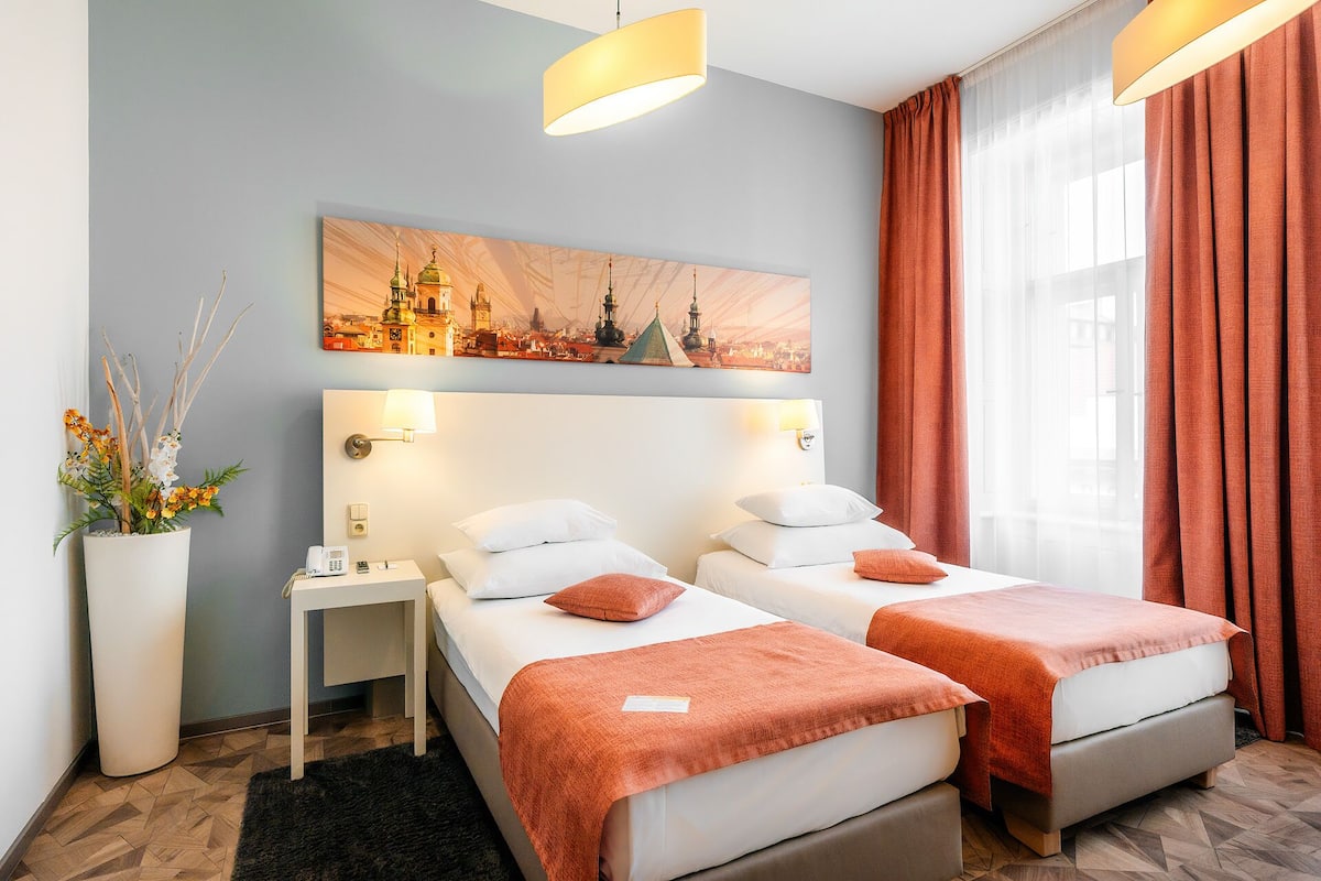 Ein Hotelzimmer mit zwei Betten, einem Nachttisch, einer Lampe, einem Stadtbild als Gemälde, einer Topfpflanze und einem Fenster mit Vorhängen.