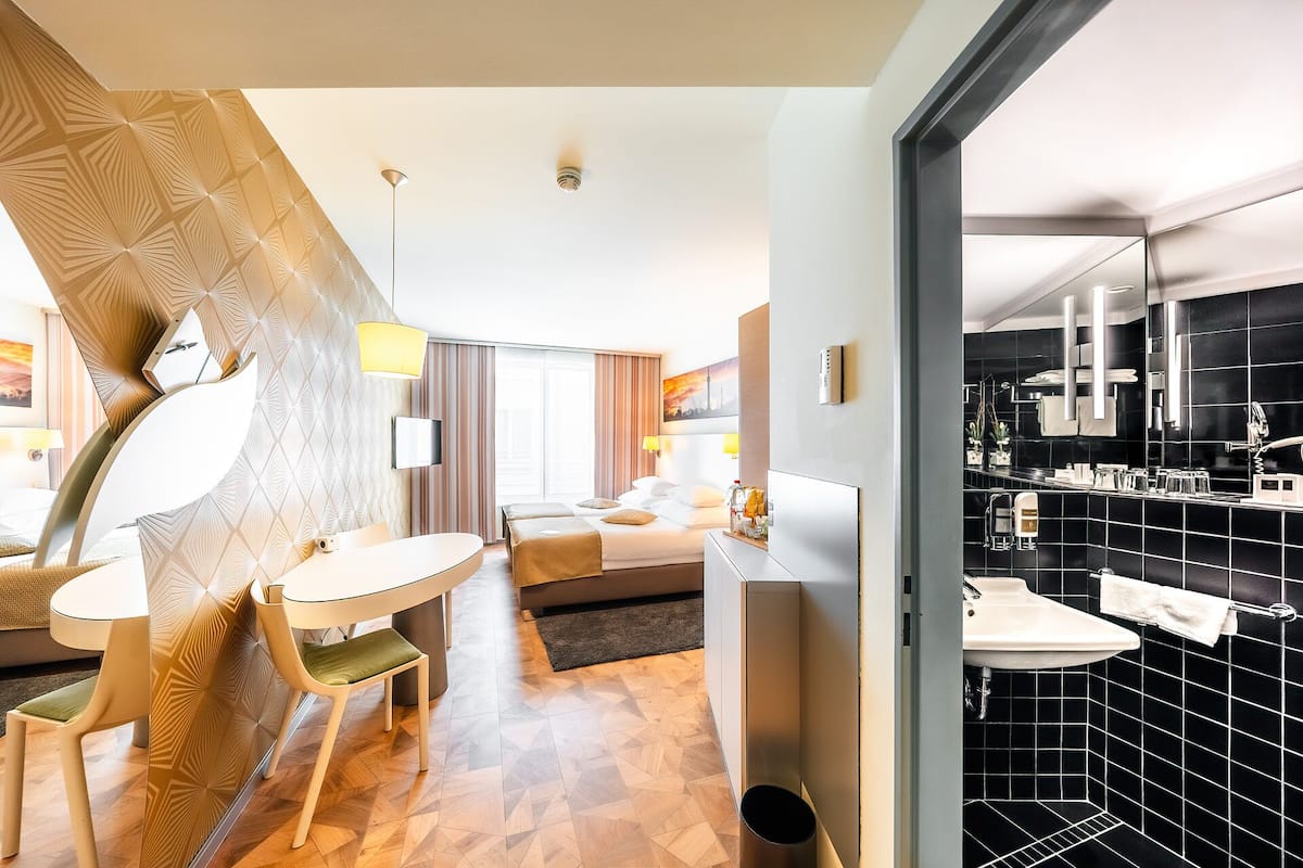 Ein Hotelzimmer mit Bett, Schreibtisch, Sessel, Fernseher und einem Badezimmer mit Waschbecken und Spiegel.