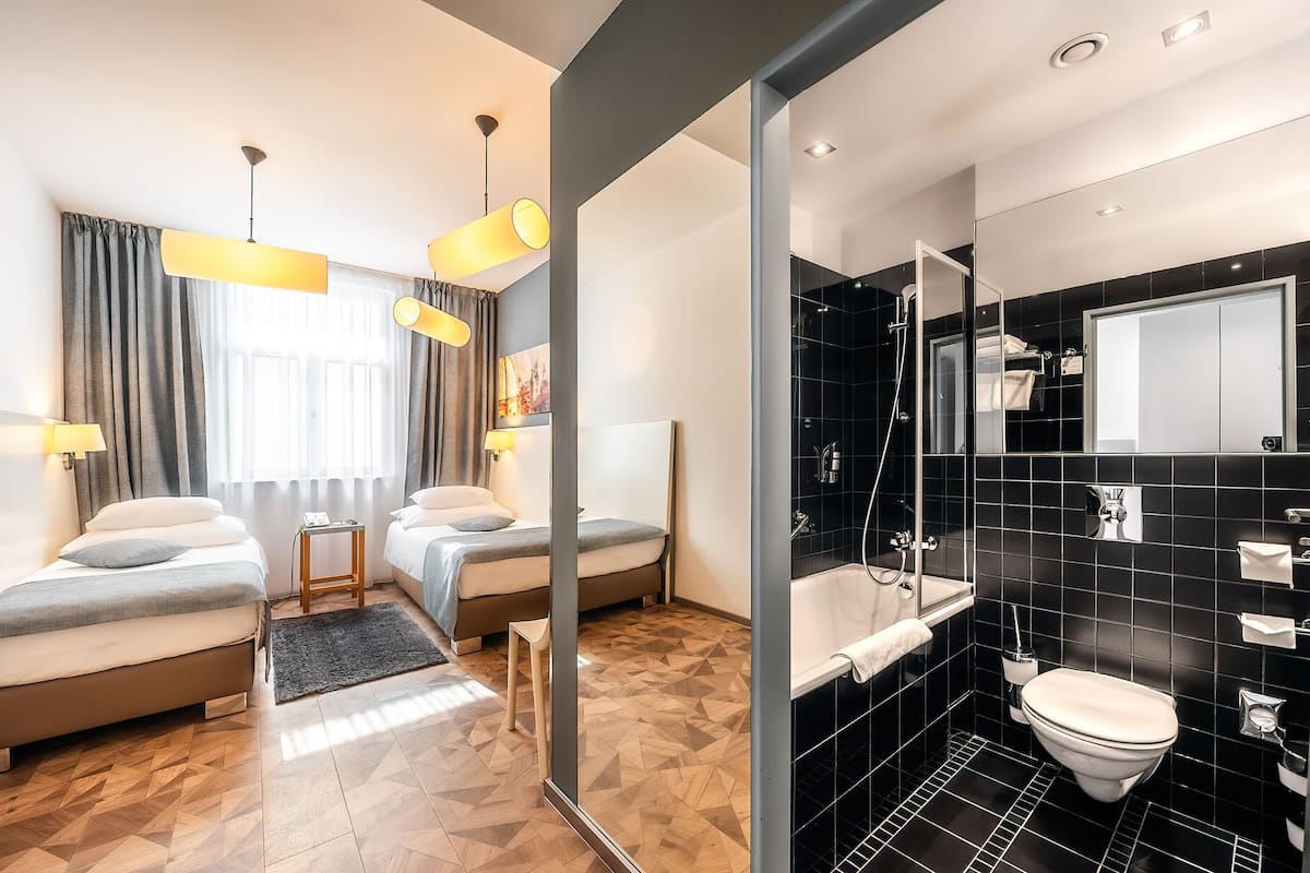 Ein Hotelzimmer mit zwei Betten, einem großen Fenster mit Vorhängen, einem Holzboden und einem modernen Badezimmer mit schwarzen Fliesen und einer Dusche.