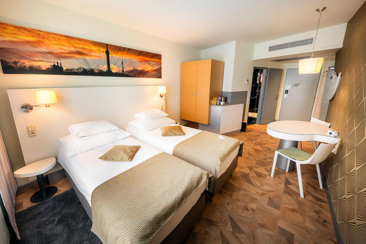 A hotel room with two beds, a desk, a chair, a small table, a painting, and a bathroom.